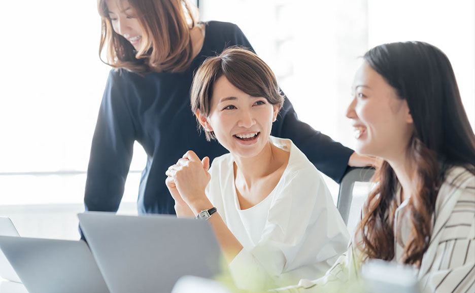 笑顔の女性3人が仕事中に会話している様子。手前にはノートパソコンが見える。