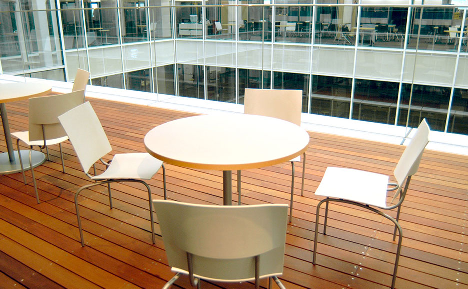 木製の床に白い丸テーブルと椅子が複数配置され、奥にはガラス張りのオフィス空間が広がる。