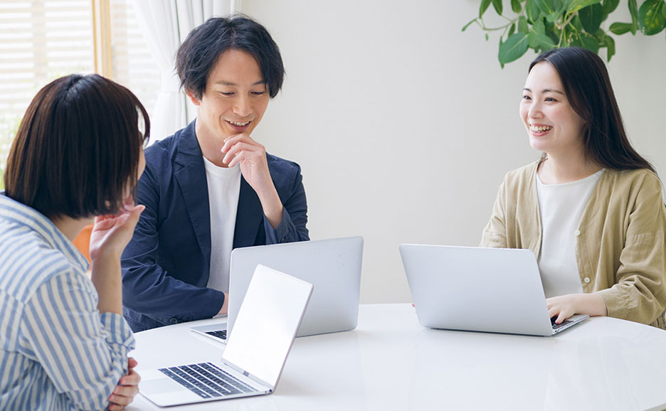 笑顔でラップトップに向かう男女3人が会議室で話し合う様子。
