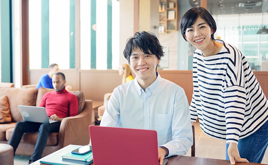 オフィスで、手前の男性は赤いノートPCを見ている。奥の女性は笑顔で男性の肩越しにカメラを見ている。