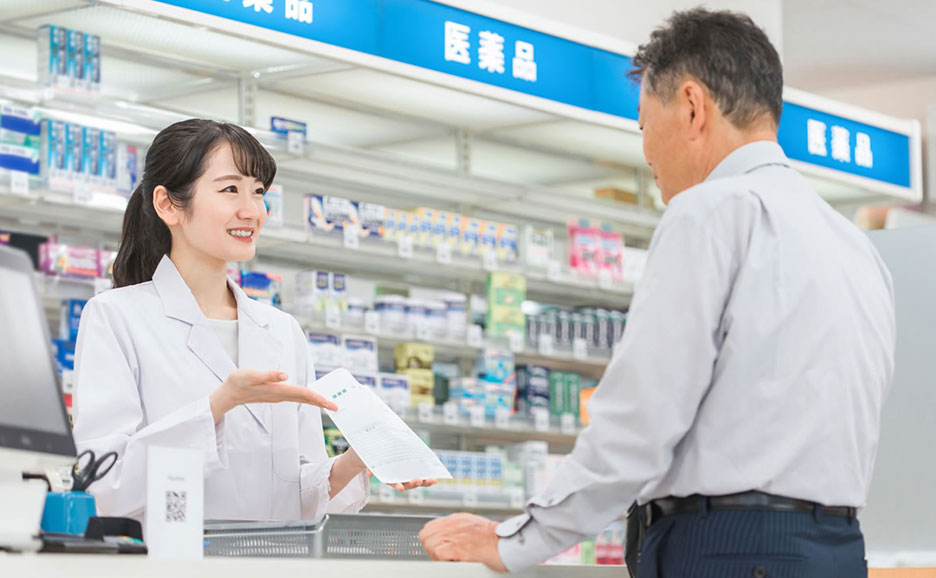 薬局で笑顔の薬剤師が書類を手に、患者と話している様子。棚には医薬品が並んでいる。