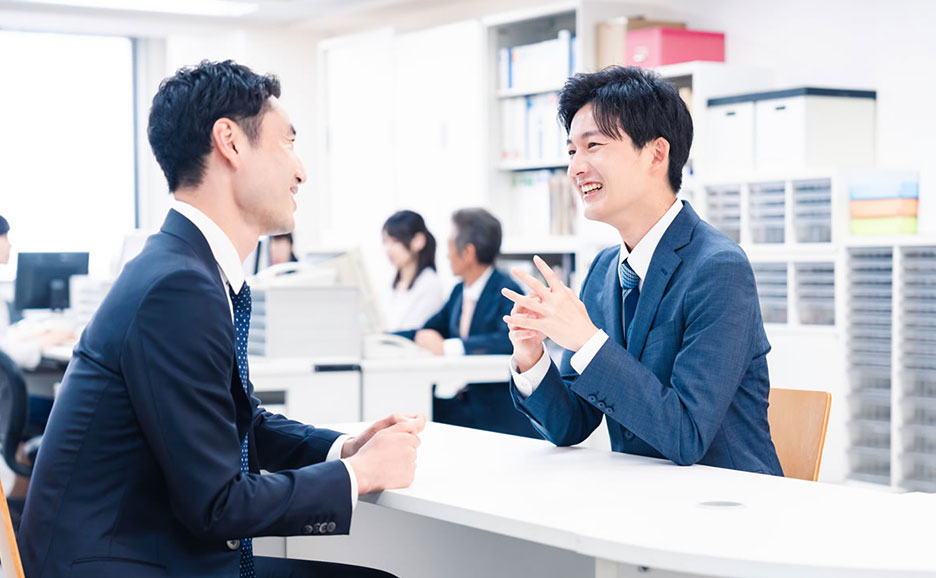 オフィスで、スーツを着た男性2人が向かい合って座り、笑顔で会話している。