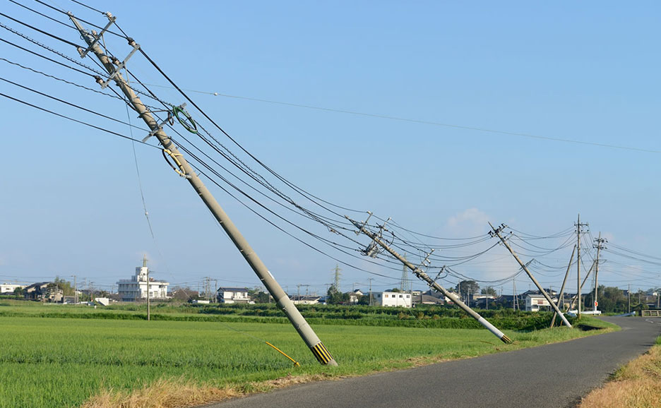 青空の下、田園風景の中に複数の電柱が傾き、電線がたるんでいる様子。アスファルトの道が手前に伸びる。