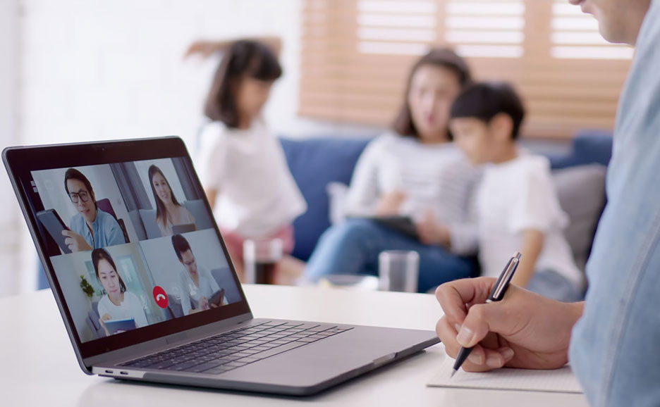 男性がノートパソコンとペンで作業中。画面にはビデオ会議の映像、背景にはソファで過ごす家族が見える。