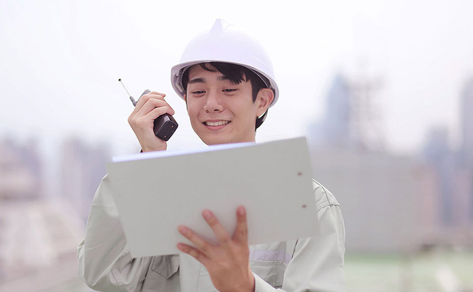 ヘルメットを被り、トランシーバーで話しながら書類を確認する若い男性作業員。笑顔。