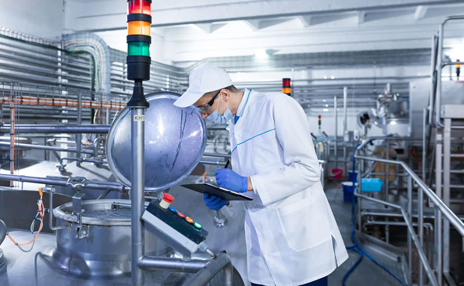 白衣を着用した作業員が、工業用タンクを点検し、クリップボードに記録を取っている様子