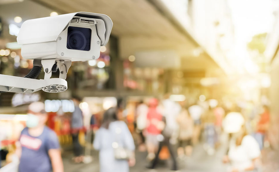 監視カメラが、明るい日差しの下、賑やかな通りと人々を見下ろしている様子。