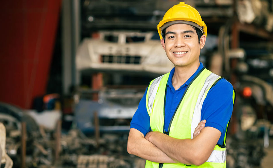 黄色いヘルメットと反射ベストを着用したアジア系の男性が、笑顔で腕を組んで立っています