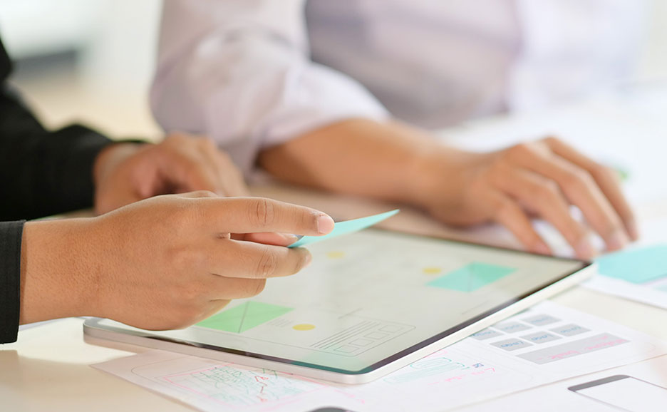 タブレットとメモ用紙が広げられたデスクで、2人の手が何かを共同で作成している様子。