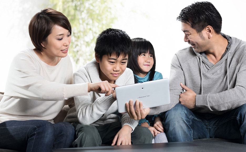 リビングルームで家族4人がソファに座り、母親が息子と一緒にタブレットを操作し、父親と娘が笑顔で見守っています。