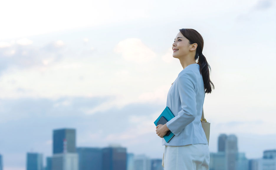 ビジネススーツ姿の女性が空を見上げ、晴れやかな表情をしています。背景はぼやけた街並み。