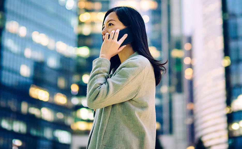 ビジネス街の夜景を背景に、スマートフォンで話しながら笑顔を見せるアジア人女性。