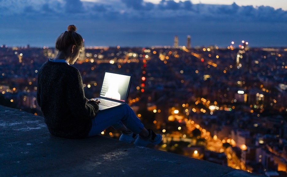 夜の街並みを見渡す屋上の縁に腰かけてノートPCを操作する女性