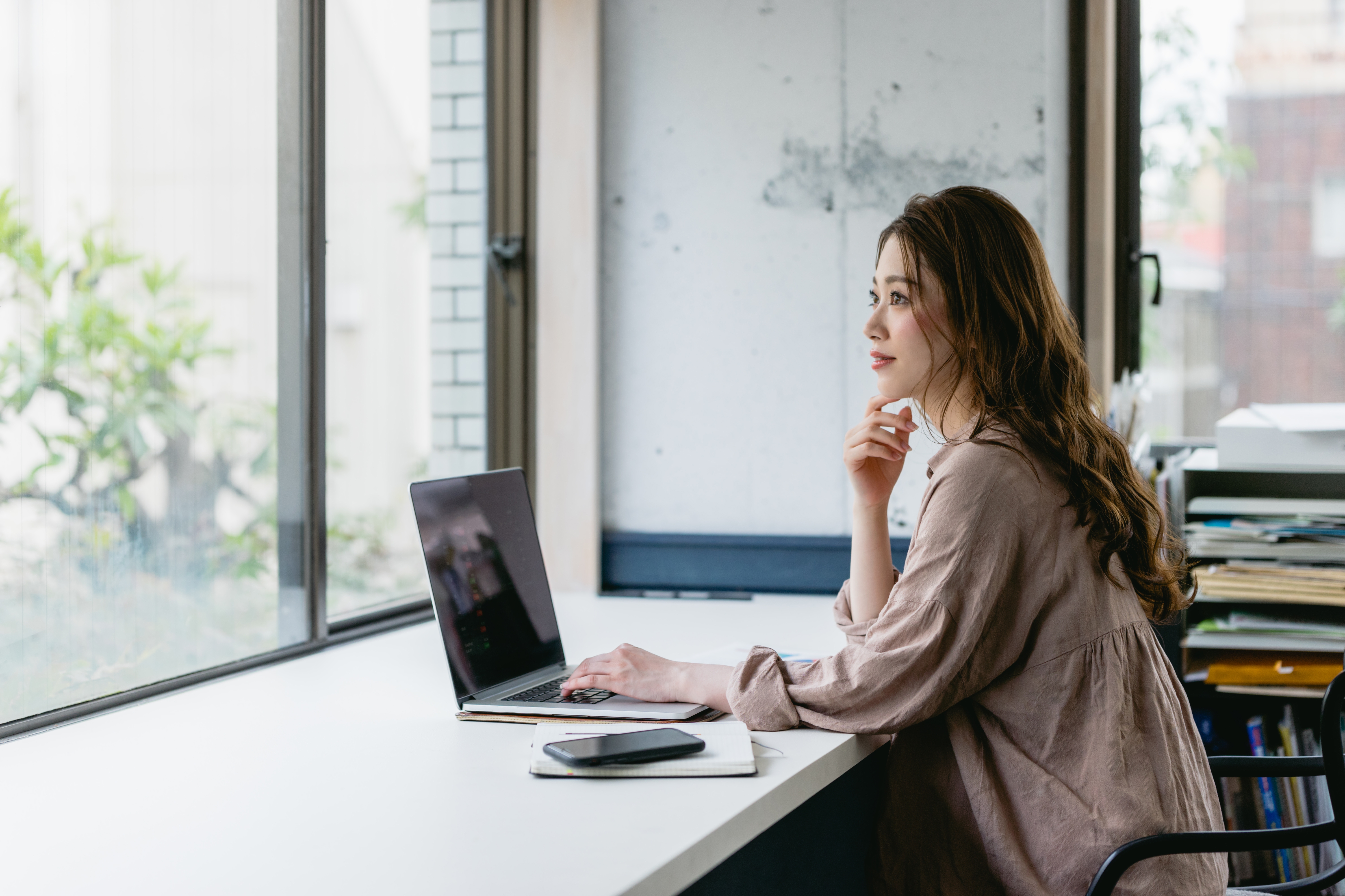 窓際の席でノートパソコンを使っている女性 窓の外を眺めている