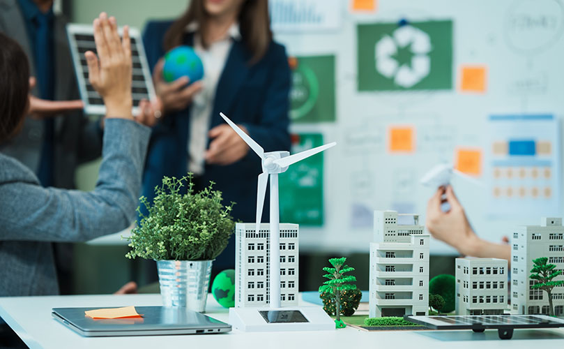 A diverse group of people discusses around a table featuring a model wind turbine, emphasizing renewable energy solutions.
