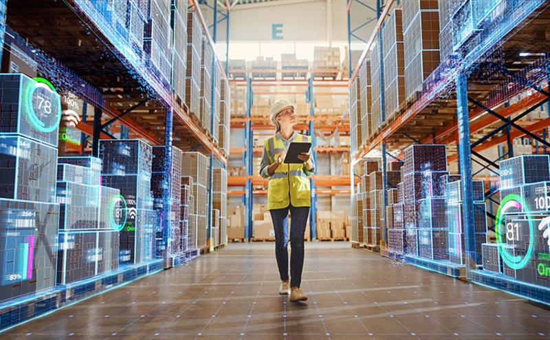 A woman stands in a warehouse, holding a tablet and looking at the screen with a focused expression.