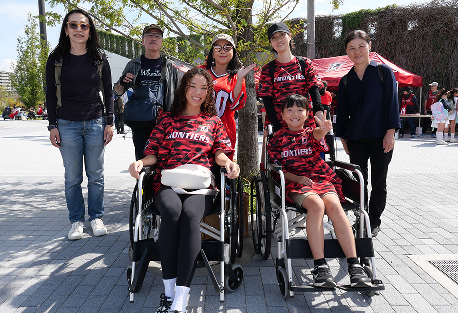 7 participants in the wheelchair experience, one of our hands-on events, are pictured in front of Fujitsu Stadium Kawasaki.2 people are pictured in wheelchairs in the center, surrounded by 5 people standing