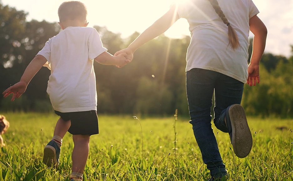 Zwei Kinder laufen Hand in Hand von hinten gesehen über eine Wiese
