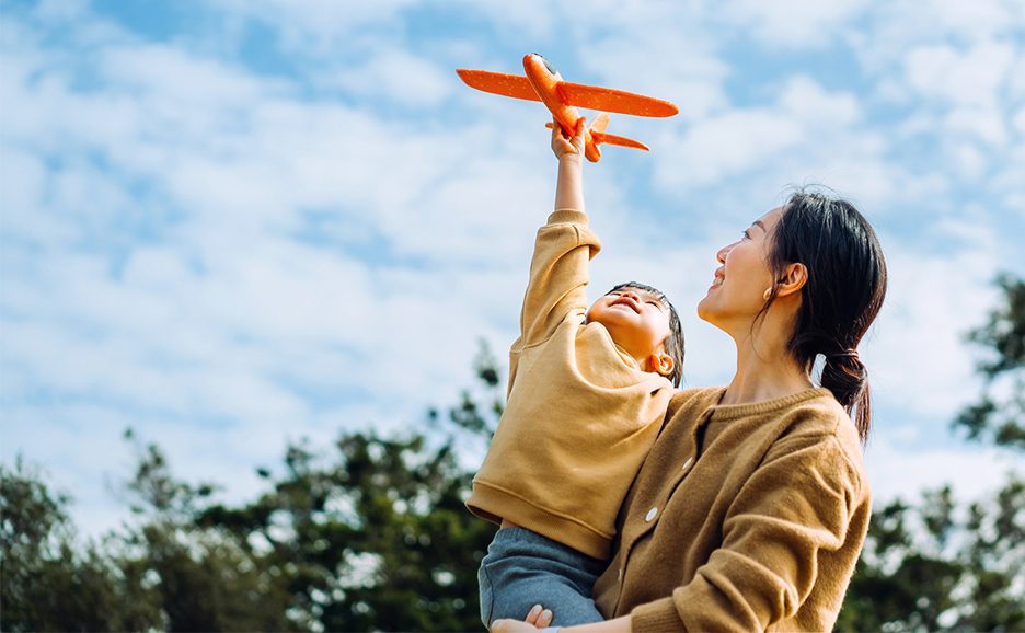 青空の下、母親が子供を抱きかかえ、子供が飛行機のおもちゃを高く掲げている写真