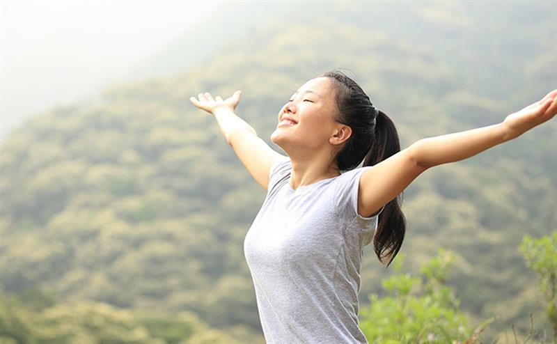A happy woman, standing in the sunshine with outstretched arms, in front of a forest