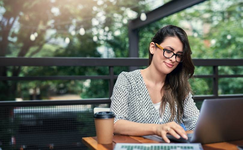 A woman working on a laptop at a terrace with green trees behind her