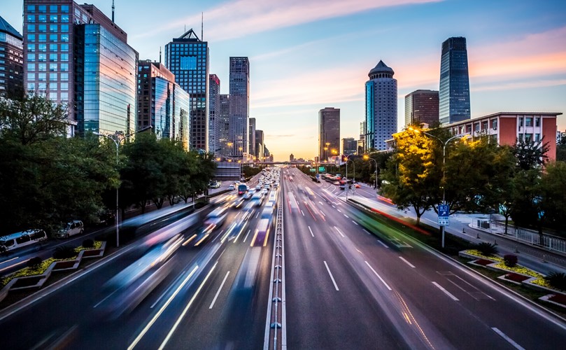  A picture of a road with vehicles in movement, surrounded by buildings.