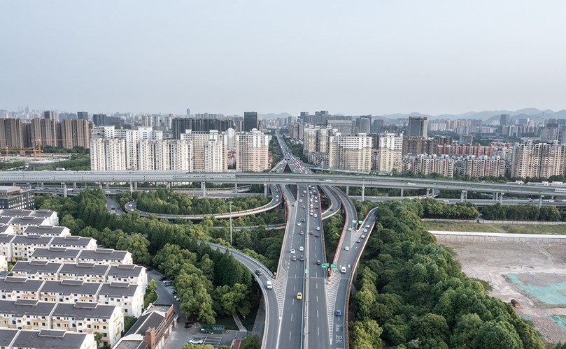 A picture with several roads and vehicles in movement, surrounded by buildings and trees.