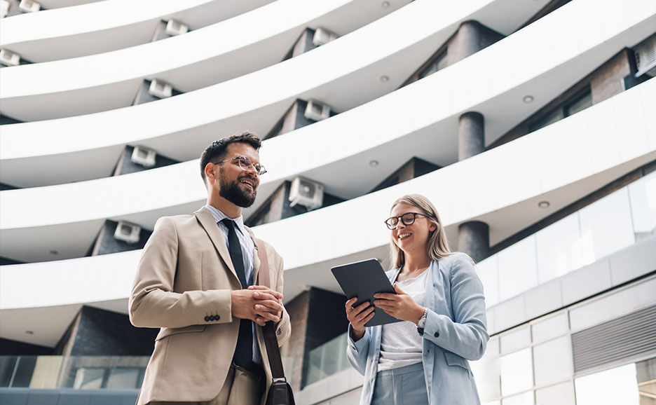 Two smiling business professionals conversing and looking at a tablet in front of a modern building.