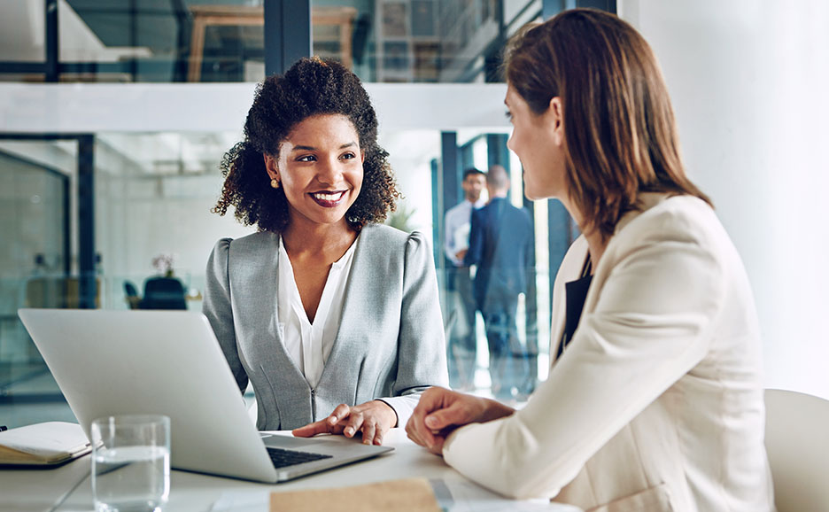 Two smiling women discussing at a laptop in an office, appearing to be consulting.