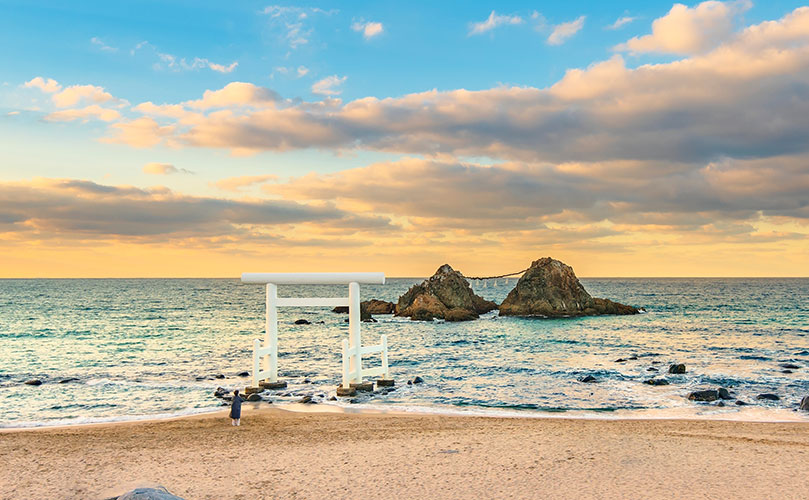 White torii gate on a calm beach, with Meoto Iwa rocks connected by a rope beyond. Beautiful Japanese coastal scene with sunset sky.