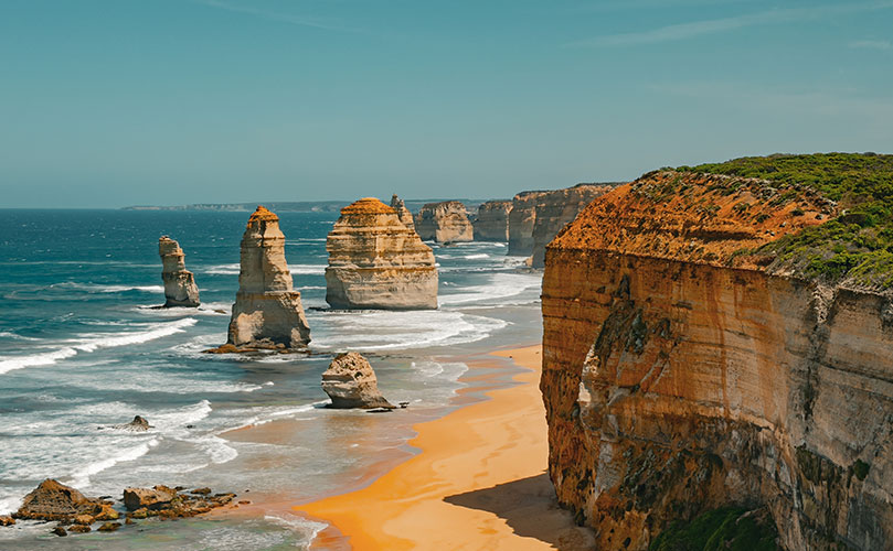 Orange beach with crashing waves and towering rock formations. Majestic coastline under a clear blue sky.