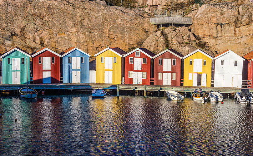 Colorful seaside boathouses reflected on calm water, with small boats moored at the dock and a rocky cliff in the background.