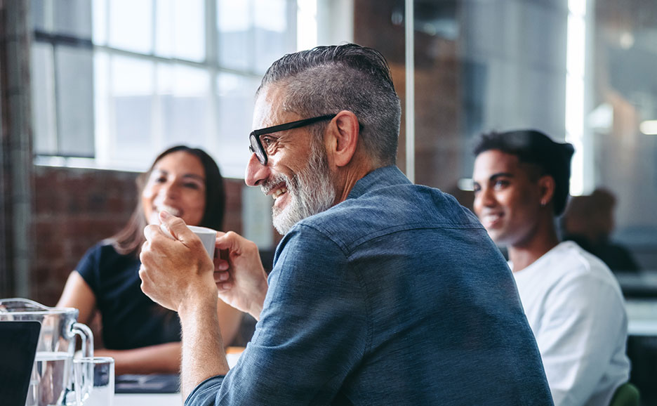 A man smiling and drinking coffee during a meeting, with diverse team members visible in the background, suggesting engaging discussion.