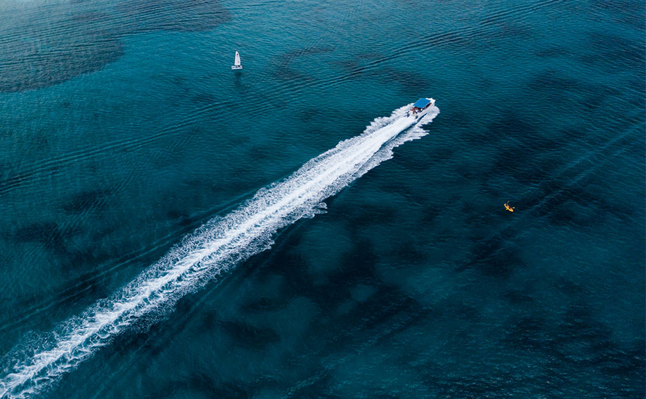 Speedboat cuts through blue water, leaving a white wake. Sailboat & kayak in distance.