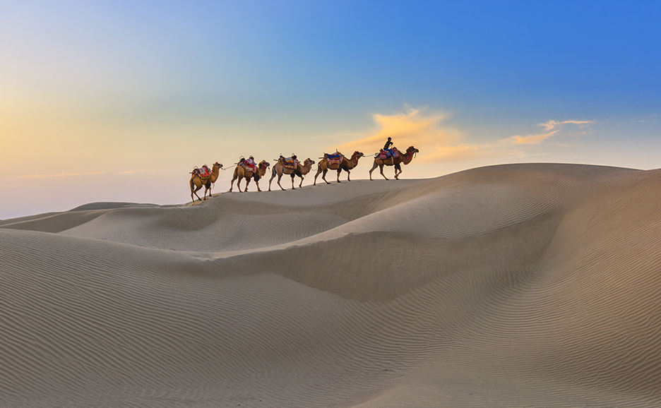 A caravan of camels carrying people across a desert at sunset.