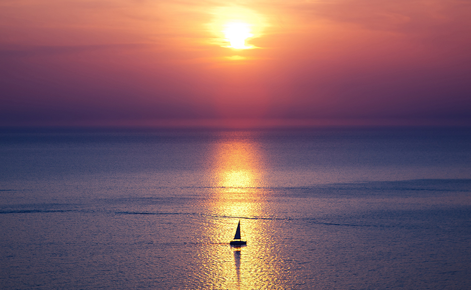 夕焼けの空と海を背景に、夕日が反射する水面を進むヨットのシルエット。