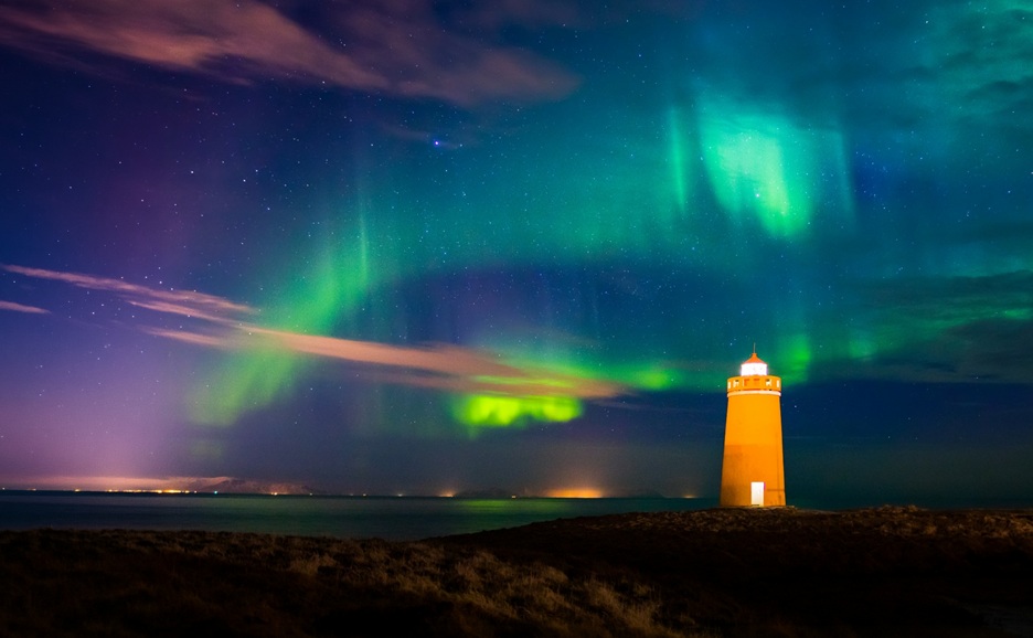 A lighthouse beam cutting through the northern lights over a calm body of water.