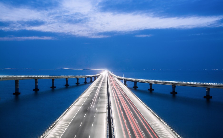 Eine lange Brücke erstreckt sich nachts über den Ozean, mit Lichtstreifen von Autos.